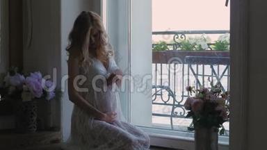 穿着白色连衣裙的孕妇正坐在窗边穿着白色蕾丝连衣裙.. 从上面看。 幸福的女人期待着